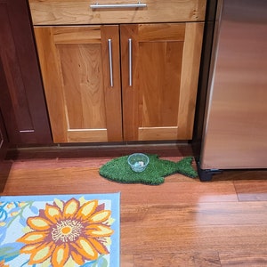 Peut inclure: Un tapis de nourriture pour animaux de compagnie en herbe artificielle verte en forme de poisson, un bol en plastique transparent avec quelques morceaux de nourriture &agrave; l'int&eacute;rieur et un tapis de sol floral bleu et orange sur un sol en bois.