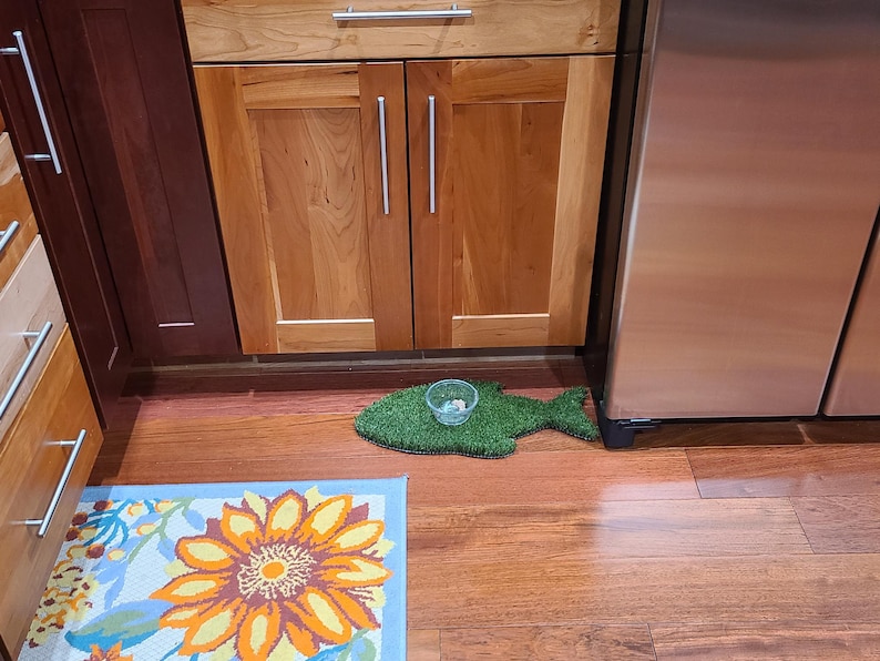 Peut inclure: Un tapis de nourriture pour animaux de compagnie en herbe artificielle verte en forme de poisson, un bol en plastique transparent avec quelques morceaux de nourriture &agrave; l'int&eacute;rieur et un tapis de sol floral bleu et orange sur un sol en bois.
