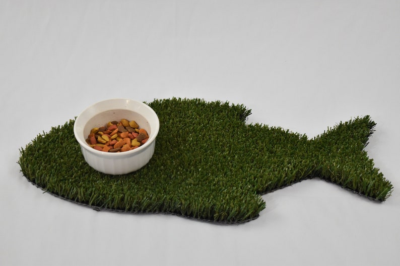 Peut inclure: Un bol en c&eacute;ramique blanche rempli de nourriture pour animaux de compagnie repose sur un tapis d'herbe artificielle verte en forme de poisson. Le tapis est con&ccedil;u pour recueillir les d&eacute;versements de nourriture et aider &agrave; garder la zone propre.