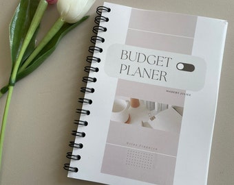 May include: A white and gray spiral bound budget planner with the text "BUDGET PLANER" and "MADE BY JULIEE". The planner has a light gray background with a white toggle switch graphic.