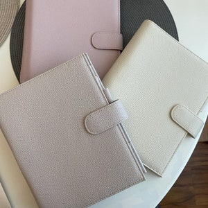 Könnte beinhalten: Drei ledergebundene Notizbücher in verschiedenen Beige-, Rosa- und Cremetönen. Jedes Notizbuch hat einen Klappverschluss mit einem Knopf.