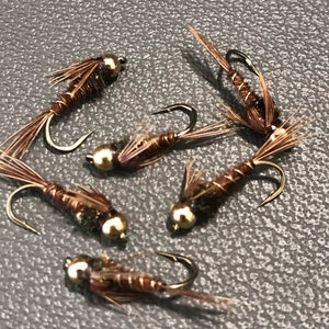 May include: Close-up of several fishing flies. Each fly has a gold-colored bead head, a brown body, and delicate feather details. The hooks are curved and metallic. The flies are arranged on a gray surface.