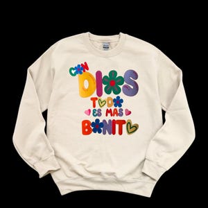 May include: A cream-colored sweatshirt with a colorful graphic design that says "Con Dios Todo Es Mas Bonito" with flowers and hearts.