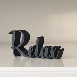 Könnte beinhalten: Dekoratives Schild mit dem Wort „Relax“ in einer stilisierten Schrift. Das Schild ist schwarz mit grauen Akzenten und steht auf einer weißen Oberfläche. Ein dekorativer Artikel.