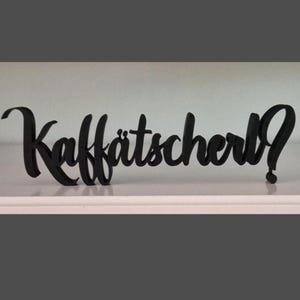 Könnte beinhalten: Ein schwarzes, dreidimensionales Dekoschild mit dem Wort "Kaffätscherl?" in einer stilisierten, verbundenen Schreibschrift. Die Buchstaben sind massiv, undurchsichtig und stehen aufrecht, was eine klare Textdarstellung ergibt.