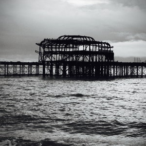 May include: A black and white photograph of a large, derelict pier stretching out into the sea. The pier is constructed from metal and features a large, open framework in the centre. The sky is overcast and the water is rough.