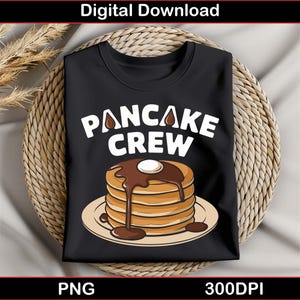 Peut inclure: T-shirt noir avec le texte "Pancake Crew" en lettres blanches. Un empilement de crêpes avec du sirop de chocolat et une cuillerée de crème fouettée est représenté sur le t-shirt.