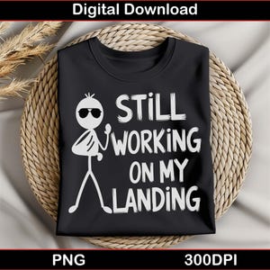 以下が含まれることがあります： 黒いTシャツに、サングラスをかけた白い棒人間と「Still Working On My Landing」という文字が書かれています。