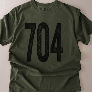 May include: Olive green t-shirt with the number "704" printed in large, distressed black font across the front. The shirt has a classic crew neck and short sleeves. The text "Made in North Carolina" is printed on the bottom right.
