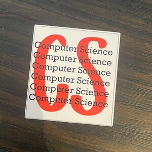 May include: A white sticker with the words "Computer Science" in red, repeated multiple times. The sticker has a large red "CS" graphic. The sticker is on a dark brown wooden surface.