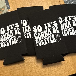 May include: Four black can coolers with the text "SO IT'S GONNA BE FOREVER" in white, along with a ring graphic. The coolers are arranged on a wooden surface, perfect for a wedding or engagement celebration.