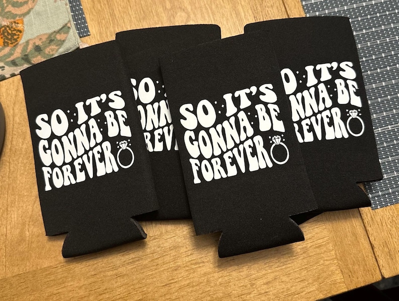 May include: Four black can coolers with the text "SO IT'S GONNA BE FOREVER" in white, along with a ring graphic. The coolers are arranged on a wooden surface, perfect for a wedding or engagement celebration.