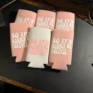 May include: A collection of pink and white can coolers with the phrase "SO IT'S GONNA BE FOREVER" printed in a retro font. The design includes a ring graphic, suggesting a wedding or engagement theme.