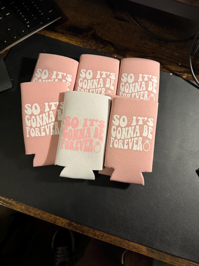 May include: A collection of pink and white can coolers with the phrase "SO IT'S GONNA BE FOREVER" printed in a retro font. The design includes a ring graphic, suggesting a wedding or engagement theme.