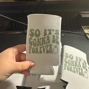 May include: A stack of light gray can coolers with the text "SO IT'S GONNA BE FOREVER" in green. The design includes a ring graphic. The coolers are made of a soft, insulating material.