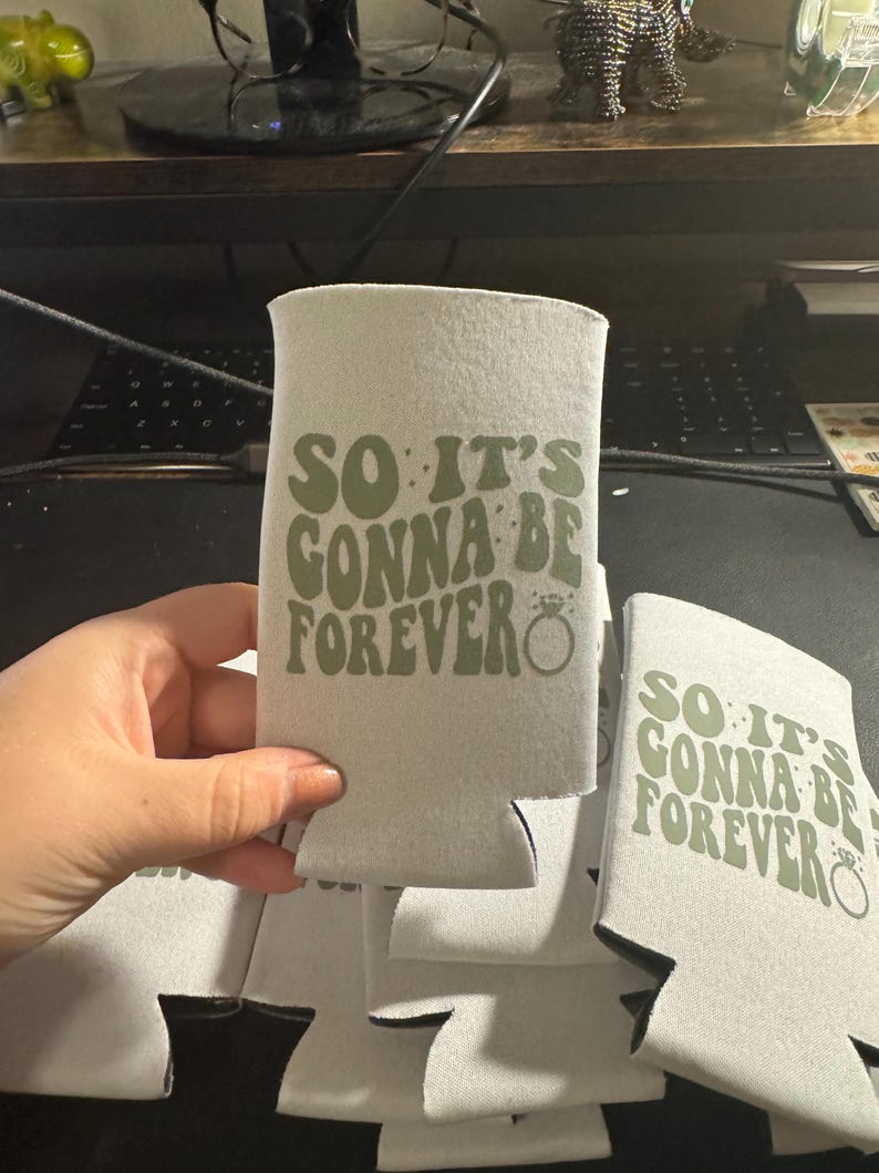 May include: A stack of light gray can coolers with the text "SO IT'S GONNA BE FOREVER" in green. The design includes a ring graphic. The coolers are made of a soft, insulating material.