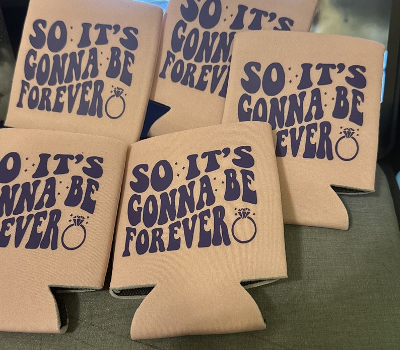 May include: Several light pink can coolers with the text "SO IT'S GONNA BE FOREVER" in dark blue, along with a diamond ring graphic. The coolers are designed to keep beverages cold and are ideal for a wedding or engagement celebration.