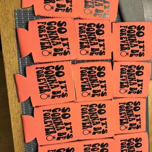 May include: A collection of orange can coolers with the text "SO IT'S GONNA BE FOREVER" printed in black. Each cooler features a ring graphic. The coolers are arranged on a wooden surface.