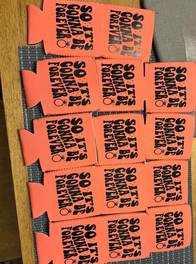May include: A collection of orange can coolers with the text "SO IT'S GONNA BE FOREVER" printed in black. Each cooler features a ring graphic. The coolers are arranged on a wooden surface.