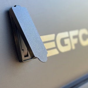 May include: A black plastic latch with a hinged cover. The latch is attached to a gray surface with the word "EGFC" printed on it.