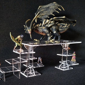 Può includere: Un set di alzatine in acrilico trasparente a cinque livelli, progettato per esporre miniature da gioco e mappe. Una miniatura di drago nero e oro è sul livello superiore, e altre miniature sono sui livelli inferiori. Il testo "RISER 5" e "3D GAME MAP EXPERIENCE" è sul lato destro dell'immagine.