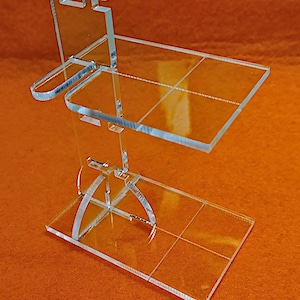 Può includere: Un supporto trasparente in acrilico con un ripiano e una base. Il supporto è progettato per contenere un piccolo oggetto, come un telefono o un tablet.
