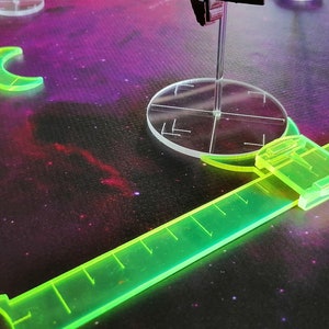 May include: A green plastic ruler with a clear plastic circle in the center. The ruler is marked with measurements and has a clear plastic handle. The circle has a crosshair in the center.