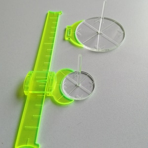 May include: A green plastic ruler with a clear plastic circle attached to it. The circle has a small clear plastic rod in the center. There is a second clear plastic circle with a rod in the center on the table.