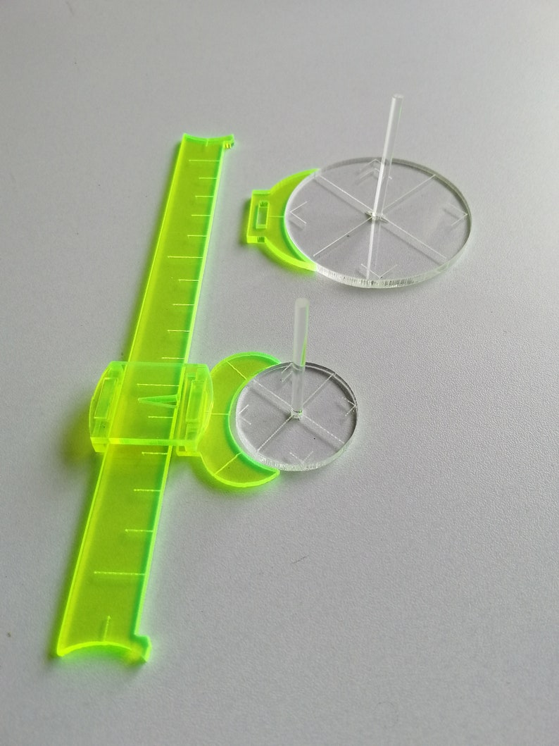 May include: A green plastic ruler with a clear plastic circle attached to it. The circle has a small clear plastic rod in the center. There is a second clear plastic circle with a rod in the center on the table.