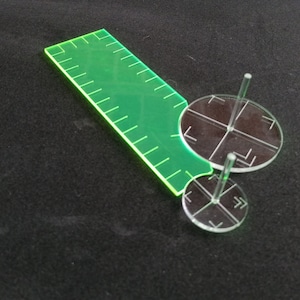 Könnte beinhalten: Ein grünes Plastiklineal mit Markierungen und drei durchsichtige Plastikkreise mit einem Mittelpfosten und Markierungen.