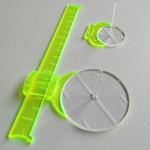 May include: Two clear plastic protractors with a green ruler. The protractors have a center point with lines radiating out to the edge of the circle. The ruler has a green handle and a clear plastic ruler with markings.
