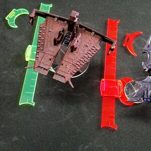 May include: Two miniature spaceships, one brown and one purple, on a black surface with clear plastic rulers. The brown spaceship has a long, thin body with a pointed nose and a large, flat wing on each side. The purple spaceship has a more rounded body with a pointed nose and a large, flat wing on each side. The spaceships are both on clear plastic bases.