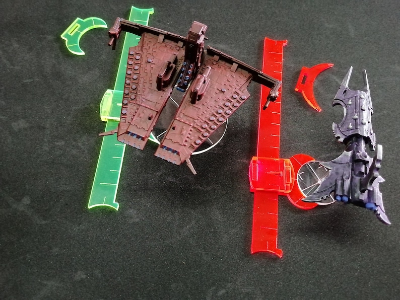 May include: Two miniature spaceships, one brown and one purple, on a black surface with clear plastic rulers. The brown spaceship has a long, thin body with a pointed nose and a large, flat wing on each side. The purple spaceship has a more rounded body with a pointed nose and a large, flat wing on each side. The spaceships are both on clear plastic bases.
