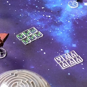 May include: A close-up of a game board with two sets of clear plastic tokens. One set has green and white symbols, and the other set has white symbols. The board is a dark purple with a galaxy pattern.
