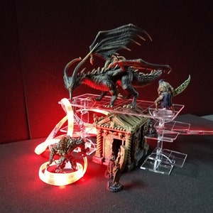 Op de afbeelding: Een zwarte drakenminiatuur met uitgespreide vleugels, een stenen gebouwminiatuur en een groen en bruin miniatuurfiguur op een transparante acrylbasis. De miniaturen staan op een zwarte ondergrond met een rode LED-lichtstrip.