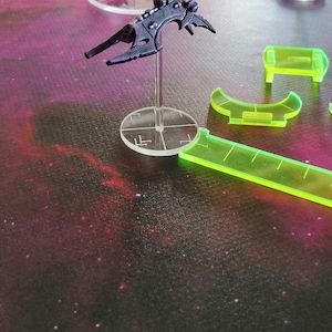 May include: A black spaceship miniature on a clear plastic base with a green ruler and green plastic measuring tools on a purple and black galaxy-themed surface.
