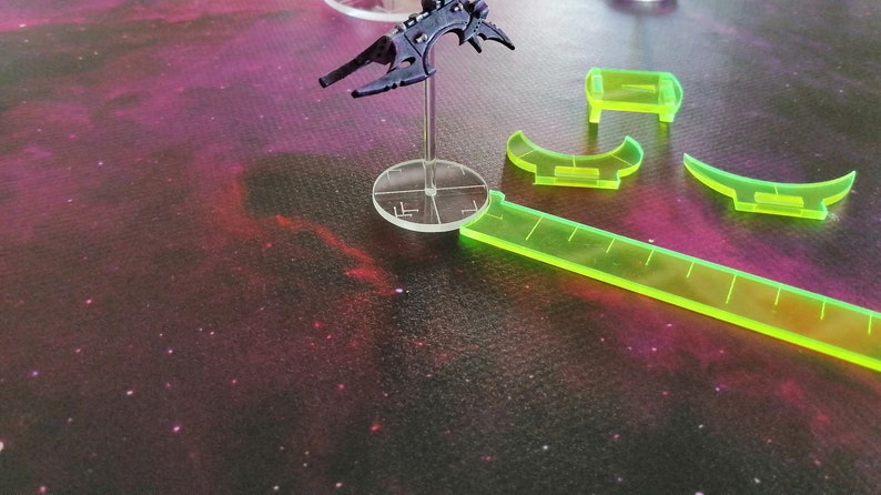 May include: A black spaceship miniature on a clear plastic base with a green ruler and green plastic measuring tools on a purple and black galaxy-themed surface.