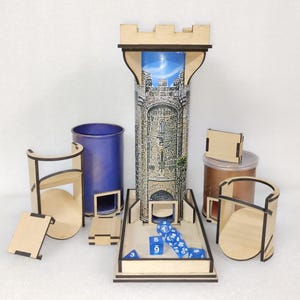 Op de afbeelding: Een houten torenvormige dobbelsteenroltoren met een kasteelbeeld aan de zijkant. De toren heeft een afneembare bovenkant en een dienblad aan de onderkant voor dobbelstenen. Er zijn ook verschillende andere houten dobbelsteenhouders en een blauwe dobbelsteenbak.