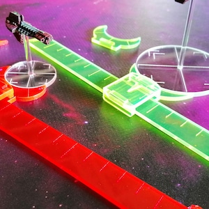 May include: A set of clear plastic rulers and a clear plastic targeting template for a tabletop game. The rulers are red and green and have markings for measuring distances. The targeting template has a clear plastic base with a crosshair design.