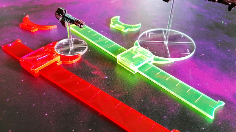 May include: A set of clear plastic rulers and a clear plastic targeting template for a tabletop game. The rulers are red and green and have markings for measuring distances. The targeting template has a clear plastic base with a crosshair design.
