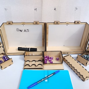 Puede incluir: Una caja de madera con dos compartimentos, cada uno con una pizarra blanca de borrado en seco. La caja tiene un pequeño cajón con compartimentos para dados. La caja está abierta y hay dados dentro. Hay un marcador negro y un bolígrafo azul sobre la mesa.