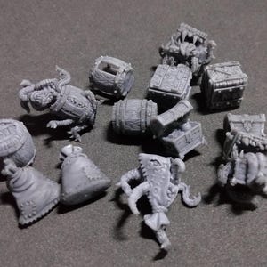 Puede incluir: Una colección de cofres del tesoro en miniatura de plástico gris, barriles y criaturas. Los cofres tienen detalles intrincados y están cerrados. Los barriles están abiertos y parecen estar hechos de madera. Las criaturas están en varias poses, algunas con la boca abierta y dientes afilados.