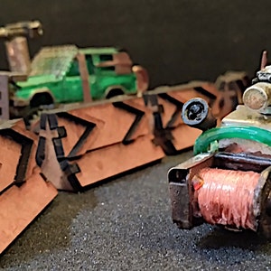 May include: A miniature, rusty, green and brown,  handmade,  toy car with a spool of pink thread and a green pipe. The car has a small, black,  gun on top. The car is in front of a row of brown, wooden,  arrow-shaped barricades.