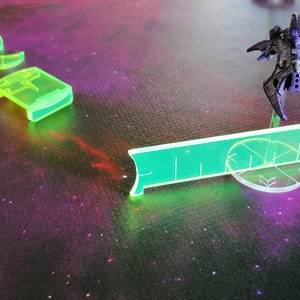 May include: A green plastic ruler with a black spaceship on a clear plastic base. The spaceship is on a white circle with a black star design. The background is a dark purple with white stars.