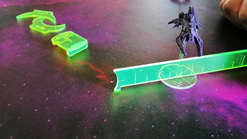 May include: A green plastic ruler with a black spaceship on a clear plastic base. The spaceship is on a white circle with a black star design. The background is a dark purple with white stars.