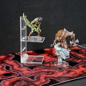 Puede incluir: Dos miniaturas de fantasía en bases de plástico transparente, una verde y otra marrón, expuestas en un soporte de plástico transparente sobre un fondo rojo y negro.