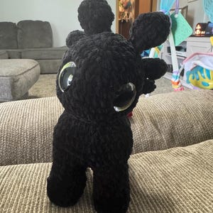 May include: A black crocheted dragon plush toy with large, reflective eyes. The plush has a rounded head, large ears, and a small, red detail near the mouth. The toy is made of a soft, textured yarn.