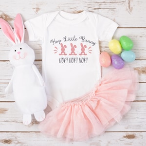 May include: White baby onesie with pink bunny silhouettes and the text "Hop Little Bunny HOP! HOP! HOP!"  A pink tutu and a white stuffed bunny with pink ears are also in the image.