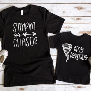 May include: Two black t-shirts with white text. The larger shirt says "Storm Chaser" with an arrow and heart. The smaller shirt says "tiny tornado" with a tornado graphic.
