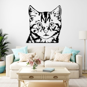 May include: A black and white cat face wall decal. The cat's face is centered, with detailed stripes and large eyes. The decal is applied to a white wall above a cream-coloured sofa with decorative cushions. A potted plant and a lamp are also visible.